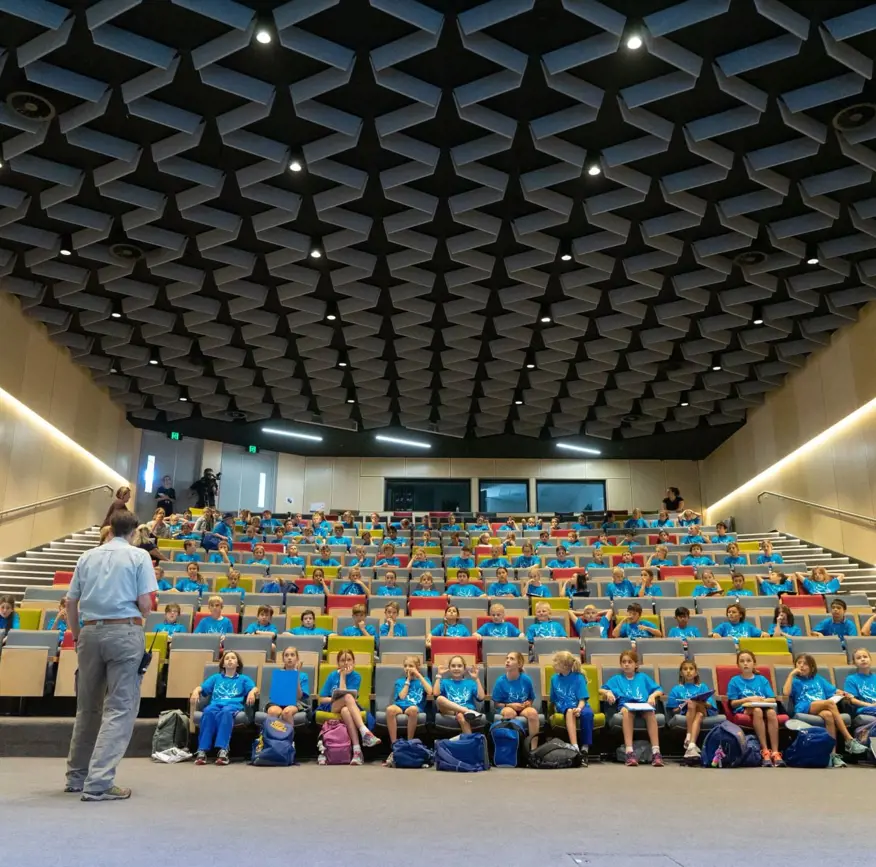 Students seated in a large auditorium listening to a teacher at the front.